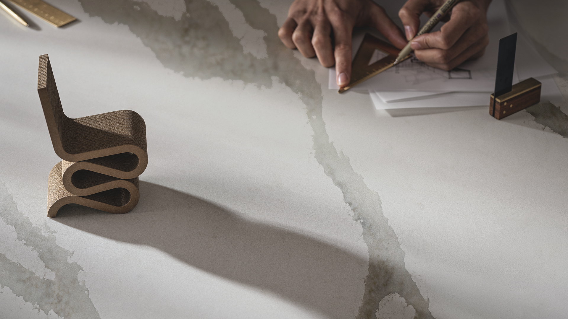 Hands sketching geometric shapes near a modern, sculptural cardboard chair model on white surface.